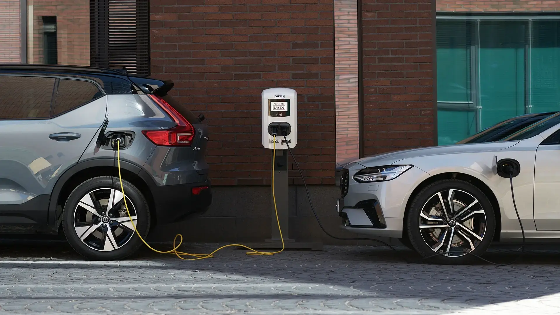 Two electric cars with AC charger in the middle with brick wall in background in city