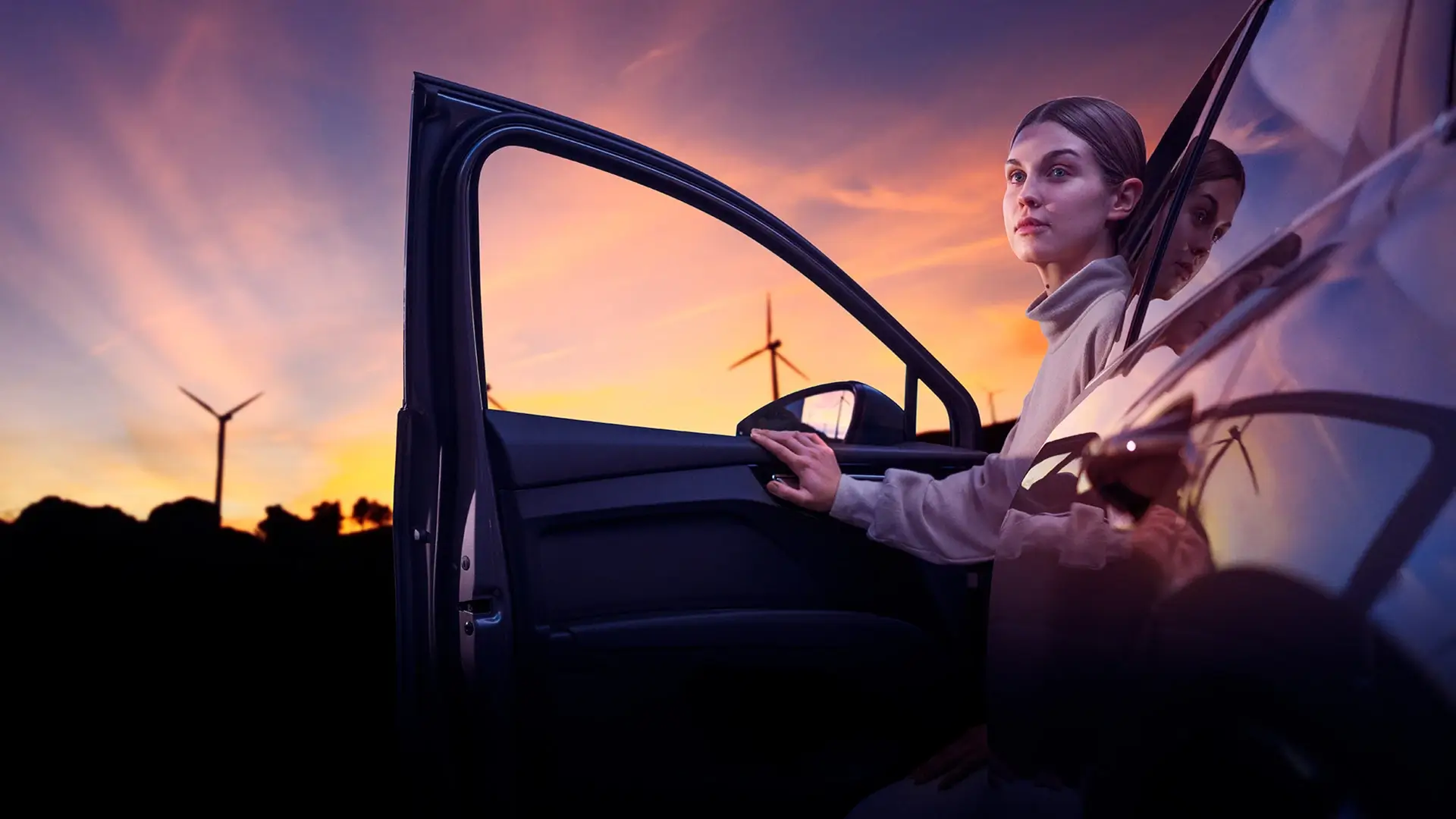 Woman sitting in electric car with wind turbines at sunset