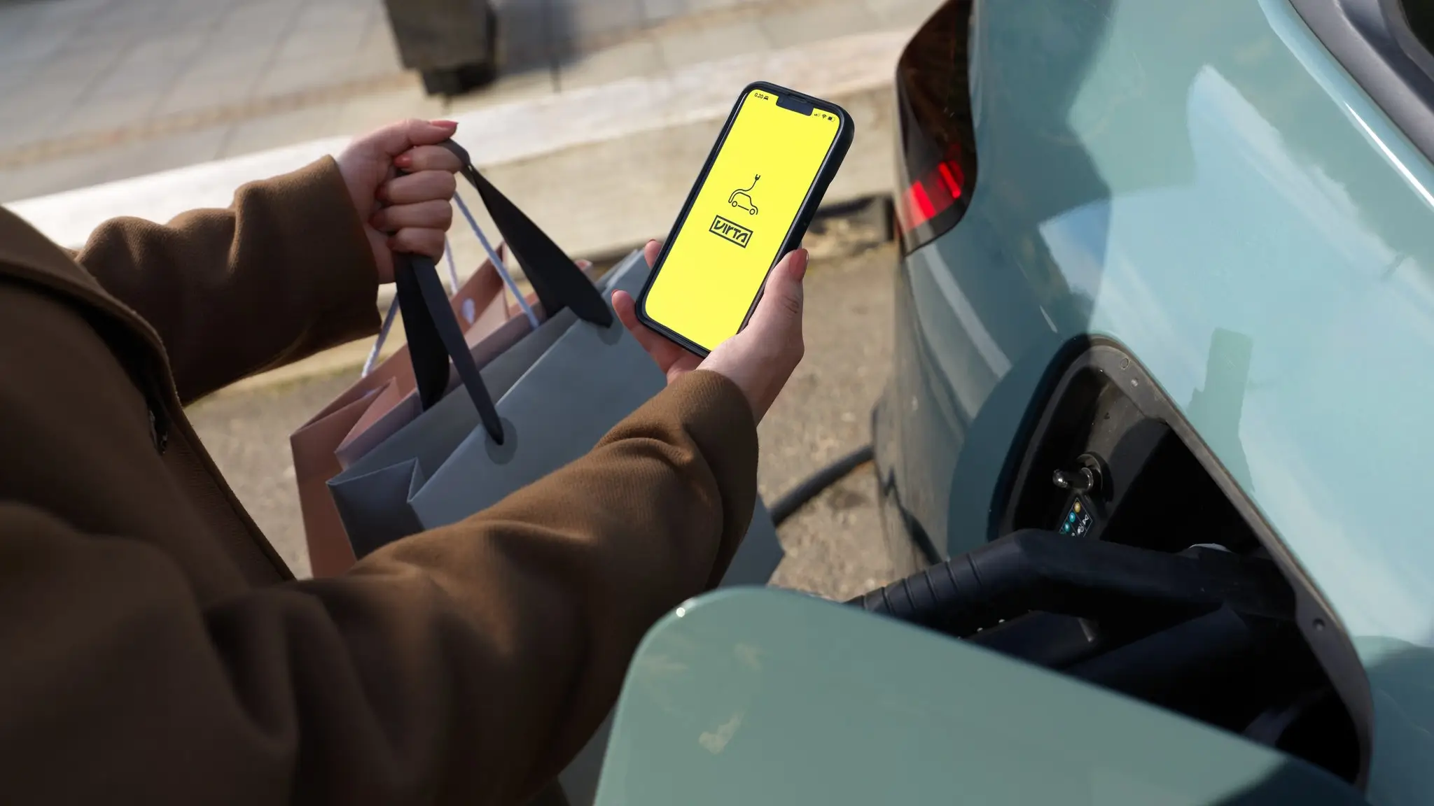 Frau mit Einkaufstüten steuert Ladevorgang am Handy während das E-Auto geladen wird